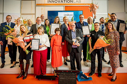 Gruppenfoto der Nominierten des Amsterdam Innovation Award 2022 auf der Messe Interclean. Die 18 Personen blicken lächelnd in die Kamera und halten Blume, Urkunden und den Award in den Händen.