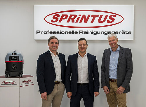 Foto der beiden Sprintus- Geschäftsführer Valentin Dahlhaus und Ermin Fazlic neben dem ehemaligen Geschäftsführer Holger Lepold. Die Männer stehen neben dem Staubsauger-Modell Era Igo von Sprintus und lächeln in die amera.