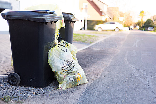 Zwei schwarze Restmülltonnen stehen am Straßenrand. Daneben liegen mehrere gefüllte gelbe Müllsäcke mit der Aufschrift "Gelber Sack"