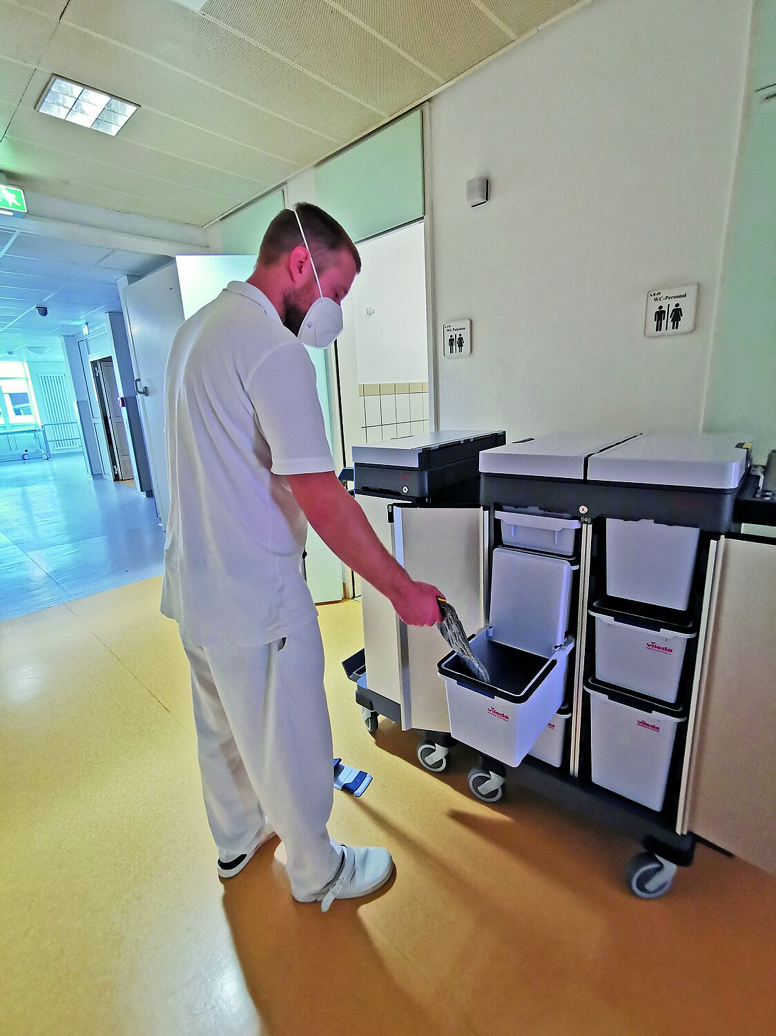 Ein Mitarbeiter in weißer Arbeitskleidung steht auf einem Flur des Dietrich-Bonhoeffer-Klinikums vor dem neuen Vileda Origo 2 Reinigungswagen. Der Wagen ist an der Vorderseite geöffnet. Im Inneren befinden sich mehrere Putzeimer, die wie Schubladen eingehängt sind. Einer der Eimer ist herausgezogen. Der Mitarbeiter steht leicht zum Wagen gebeugt und legt einen Flachwischer-Aufsatz in den herausgezogenen Eimer.