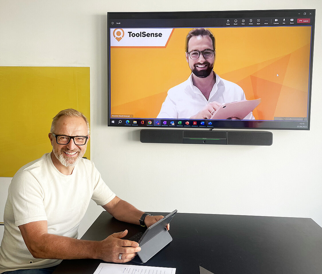 Markus Sontheimer, CIDO von ISS A/S, sitzt an einem Konferenztisch und lächelt in die Kamera, während er ein Tablet in der Hand hält. Über ihm hängt ein Bildschirm, auf dem Alexander Manafi, CEO von ToolSense in einem Videocall-Fenster zu sehen ist und ebenfalls ein Tablet in der Hand hält und in die Kamera lächelt.