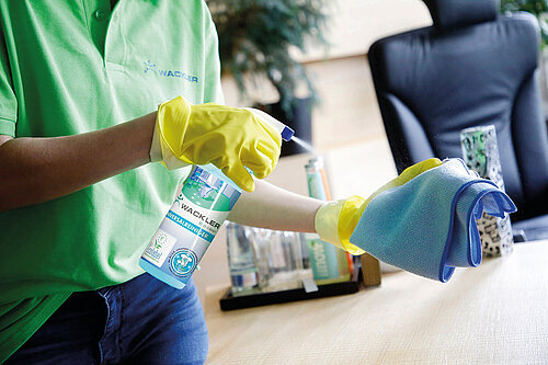 Nahansicht der Hände einer Reinigungskraft mit gelben Handschuhen. Die rechte Hand hält eine durchsichtige Sprühflasche mit der Aufschrift "Wackler Universalreiniger" und dem Ecolabel auf dem Etikett, mit der sie auf einen blauen Putzlappen in der linken Hand sprüht. Die Person trägt ein türkisfarbenes Polo-Shirt mit der Aufschrift "Wackler". Im Hintergrund ist ein Konferenzraum zu sehen.