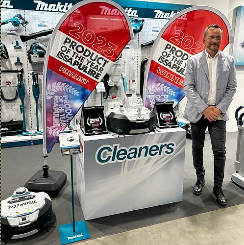 Stefano Mattsini, Marketingleiter bei der Makita SpA am Makita-Messestand neben dem Reinigungsroboter DRC300Z. Der Roboter steht auf einem Podest mit der Aufschrift "Cleaners". Links und rechts neben dem Podest stehen Banner mit der Aufschrift "2023. Product of the Year. ISSA Pulire. Finalist." Im Hintergrund sind weitere Reinigungsgeräte von Makita ausgestellt.