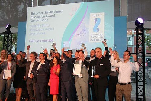 Gruppenfoto der Gewinner des Purus Innovation Award mit der Jury auf der Bühne auf der CMS Berlin. Die 21 Personen stehen jubelnd vor einer Leinwand mit der Aufschrift "Besuchen Sie die Purus Innovation Award Sonderfläche. Hall 5.2, Stand 100."