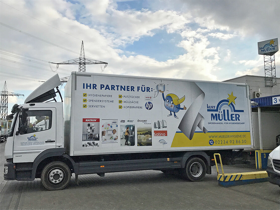 Ein weißer LKW steht rückwärts an einer Laderampe. Auf dem LKW ist das Logo von Kurt Müller zu sehen sowie der Text: "Ihr Partner für: Hygienepapiere, Putztücher, Spendersysteme, Müllsäcke, Servietten, Kopierpapiere. www.mueller-hygiene.de. Telefon: 02234928630" Außerdem sind die Logos von Xerox, HP, Katrin, Wepa, Tork, Lucart, Satino und WBV auf dem LKW abgebildet. Im Hintergrund ist ein Schild mit dem Logo von Kurt Müller zu sehen.