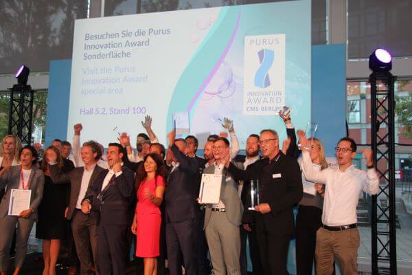 Gruppenfoto der Gewinner des Purus Innovation Award mit der Jury auf der Bühne auf der CMS Berlin. Die 21 Personen stehen jubelnd vor einer Leinwand mit der Aufschrift "Besuchen Sie die Purus Innovation Award Sonderfläche. Hall 5.2, Stand 100."