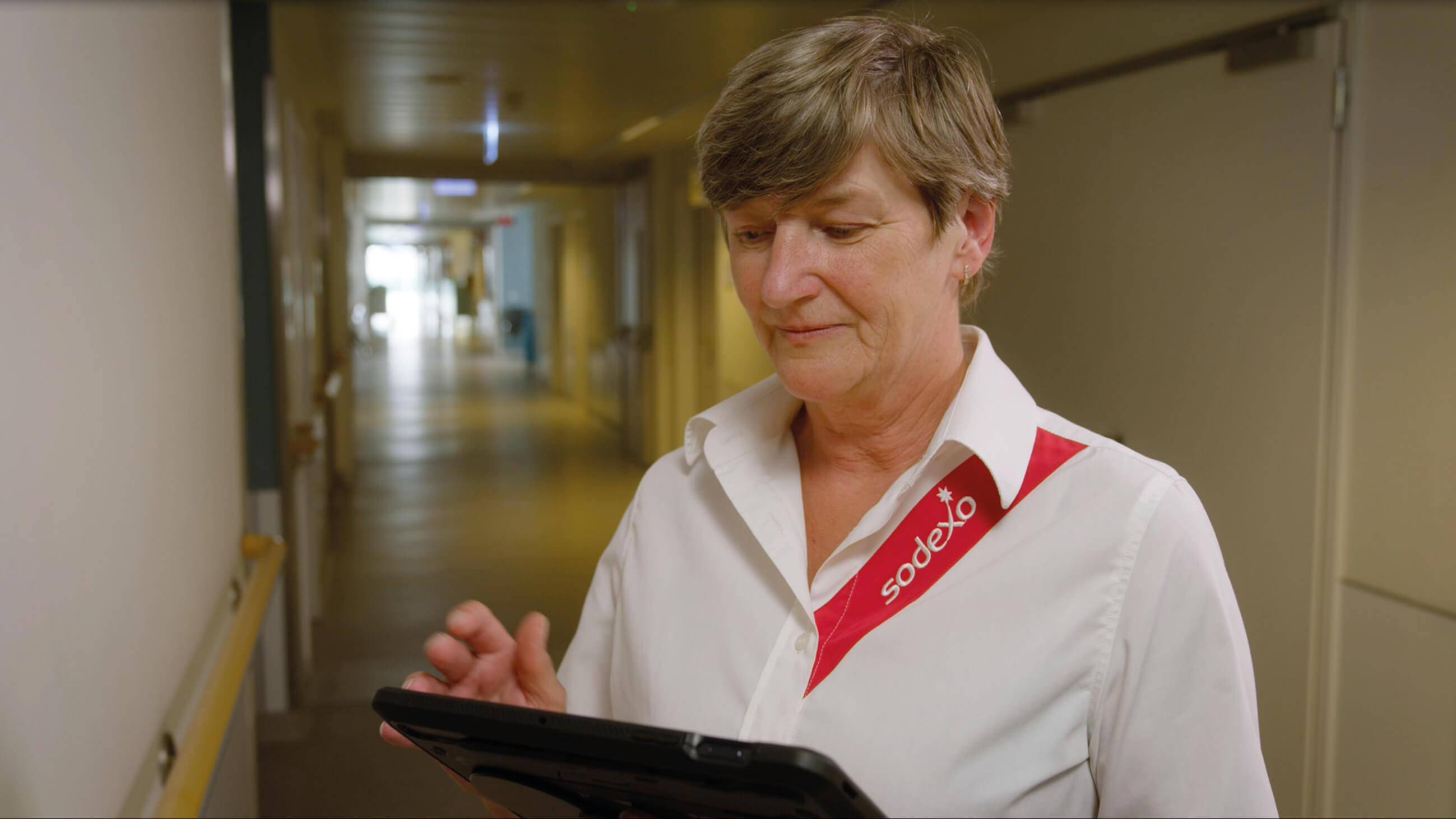Eine Frau in weißer Arbeitskleidung mit Sodexo-Aufschrift steht auf einem Krankenhausflur und blickt auf ein Tablet, das sie in der Hand hält