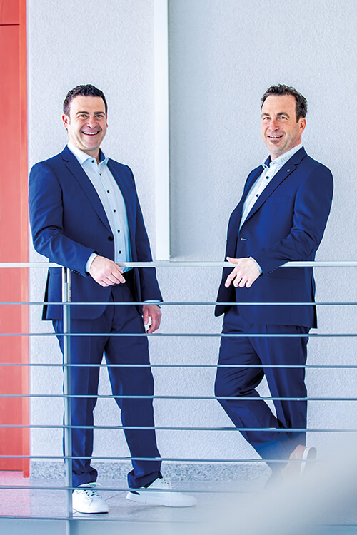 Martin und Andreas Lutz, Geschäftsführer des Forschungs- und Prüfinstituts für Facility Management GmbH, lehnen locker an einem Geländer und lächeln in die Kamera