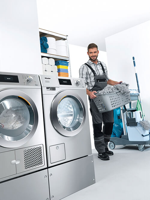 Ein Mann in Arbeitskleidung, der einen Wäschekorb mit Reinigungstextilien zu einer Waschmaschine und einem Wäschetrockner aus der Mopstar-Serie von Miele trägt. Im Hintergrund ist ein Reinigungswagen und ein Regal mit Putzutensilien zu sehen.